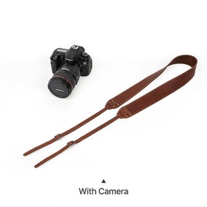 May include: A brown leather camera strap with metal buckles and stitching. A black Canon camera with a lens is in the background. The text "With Camera" is at the bottom of the image.