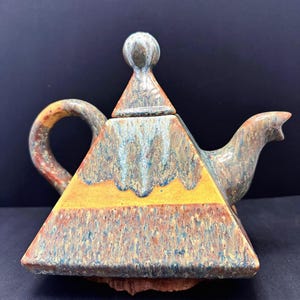May include: A handcrafted ceramic teapot with a pyramid shape. The teapot features a handle, spout, and lid with a spherical knob. The color scheme includes shades of yellow, brown, blue, and orange, creating a unique, artistic appearance.