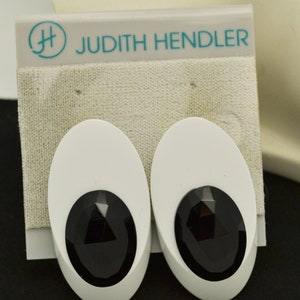 May include: A pair of white oval-shaped earrings with black faceted stones. The earrings are on a white card with the text "Judith Hendler" and a logo.