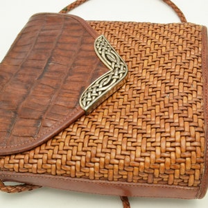 Puede incluir: Bolso bandolera de cuero marr&oacute;n con textura tejida en el cuerpo principal y solapa con relieve de cocodrilo. El bolso presenta una pieza decorativa plateada en la esquina y una correa trenzada.