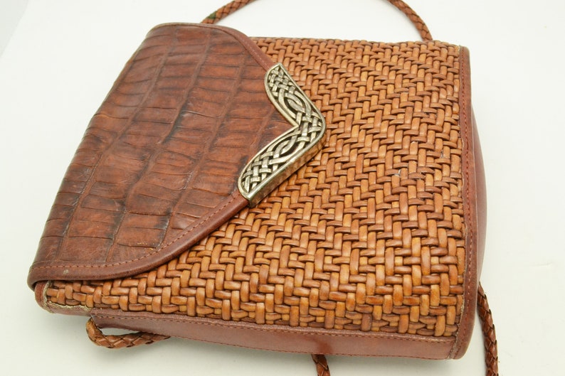 Puede incluir: Bolso bandolera de cuero marr&oacute;n con textura tejida en el cuerpo principal y solapa con relieve de cocodrilo. El bolso presenta una pieza decorativa plateada en la esquina y una correa trenzada.
