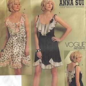 May include: A Vogue Patterns sewing pattern featuring two Anna Sui designed dresses. One dress is black with white polka dot trim and ruffles. The other dress is white with a floral print and a tie waist. The pattern includes the text "ANNA SUI" and "VOGUE PATTERNS."