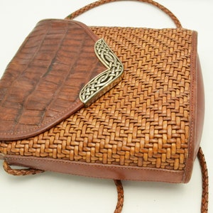 Puede incluir: Un bolso de mano de cuero marr&oacute;n con un dise&ntilde;o tejido y cierre de solapa. La solapa tiene una textura de cocodrilo y un elemento decorativo plateado. El bolso tiene una correa trenzada.