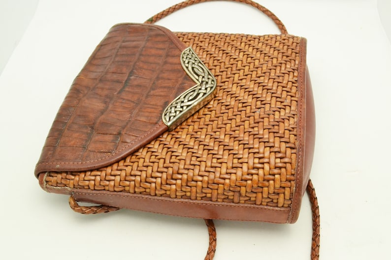 Puede incluir: Un bolso de mano de cuero marr&oacute;n con un dise&ntilde;o tejido y cierre de solapa. La solapa tiene una textura de cocodrilo y un elemento decorativo plateado. El bolso tiene una correa trenzada.