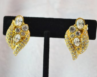 May include: A pair of gold-toned clip-on earrings. Each earring features a leaf-like design, encrusted with clear, sparkling rhinestones. The earrings have an oval-shaped, clear stone at the top and bottom, with smaller stones in between.