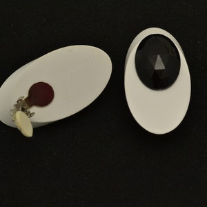 May include: A pair of white oval-shaped clip-on earrings with black gemstone accents. One earring has a black gemstone with a triangular facet, the other has a red backing.