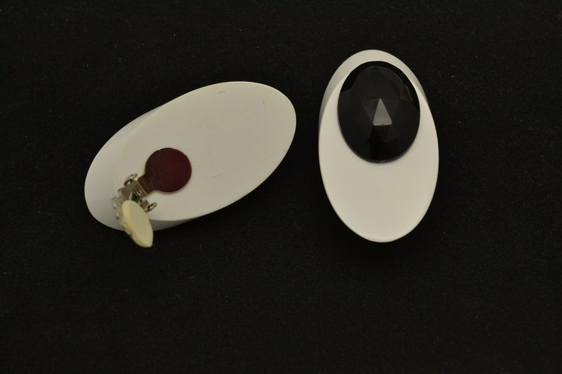 May include: A pair of white oval-shaped clip-on earrings with black gemstone accents. One earring has a black gemstone with a triangular facet, the other has a red backing.