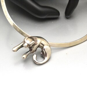 May include: A silver bangle bracelet featuring a silver cat charm. The cat is sitting on its haunches with its tail curled around its body.