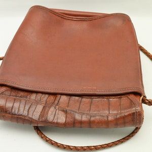 Puede incluir: Un bolso de mano de cuero marr&oacute;n con cierre de solapa y una correa trenzada para el hombro. La parte inferior del bolso presenta un patr&oacute;n texturizado similar al de un cocodrilo. El bolso es de cuero y tiene un aspecto vintage.