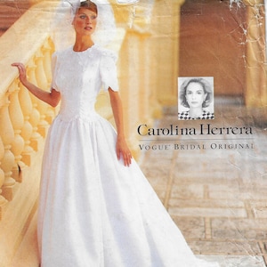 May include: A white wedding dress with short sleeves, a fitted bodice, and a full skirt, featuring a veil and floral embellishments. The text reads Carolina Herrera Vogue Bridal Original, accompanied by a small portrait of a woman in a checkered top.