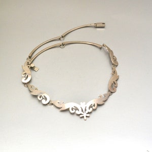 May include: A silver necklace with a delicate, intricate design. The necklace is made up of a series of connected, curved shapes that resemble leaves or flowers. The center of the necklace features a larger, more elaborate design.