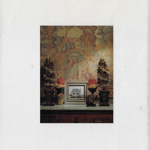 May include: A small framed picture of a house with a white exterior and a green roof. The picture is on a table with two small potted plants and a small rabbit figurine. The background is a tapestry with a forest scene.