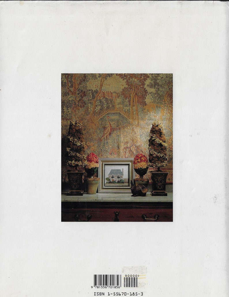 May include: A small framed picture of a house with a white exterior and a green roof. The picture is on a table with two small potted plants and a small rabbit figurine. The background is a tapestry with a forest scene.