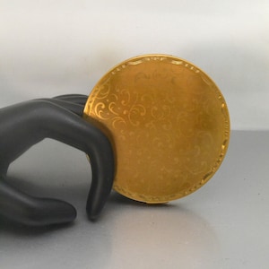 May include: A gold-colored round compact with a floral design. The compact is slightly open and appears to be made of metal.