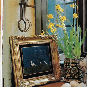 May include: A framed needlepoint picture of white geese under a crescent moon against a dark blue background. The frame is gold and ornate. A vase of yellow daffodils sits on a wooden table next to the frame.