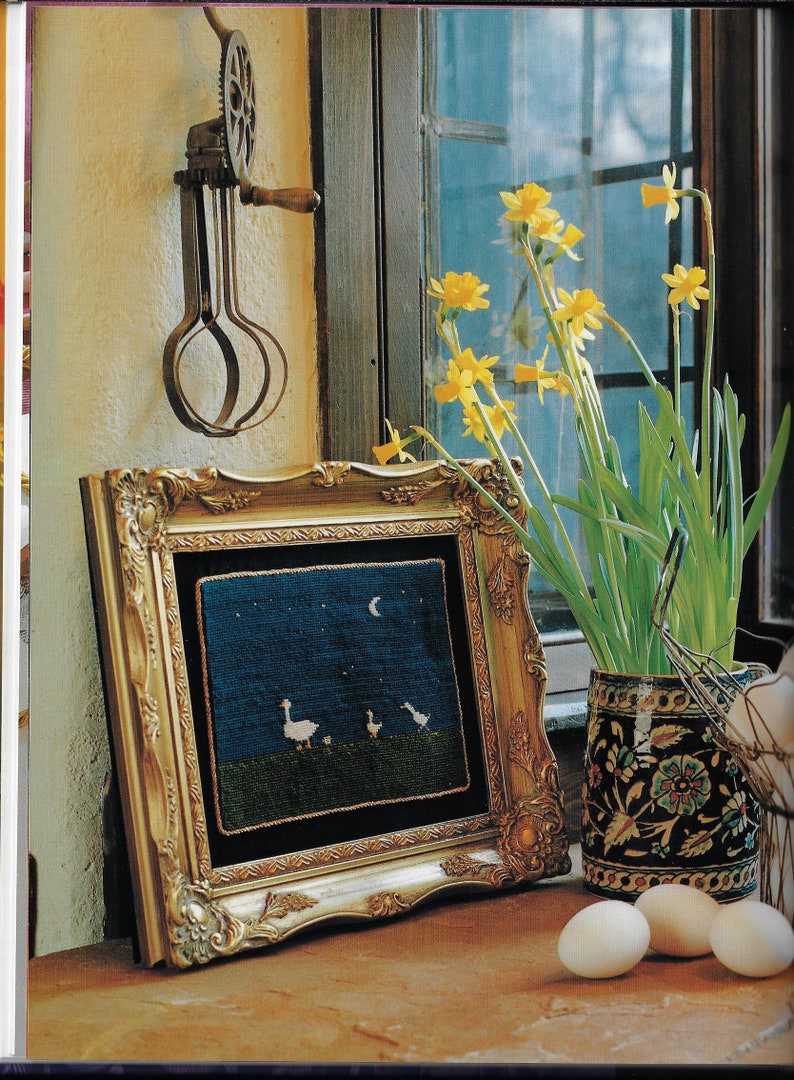 May include: A framed needlepoint picture of white geese under a crescent moon against a dark blue background. The frame is gold and ornate. A vase of yellow daffodils sits on a wooden table next to the frame.