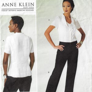 May include: A white blouse with a V-neck and short sleeves. The blouse has a bow detail at the neckline. The model is wearing black pants. The text "Anne Klein New York Vogue Patterns American Designer" is at the top of the image.