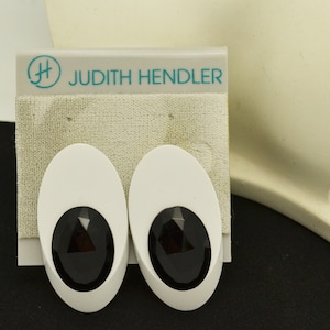 May include: A pair of white oval-shaped earrings with black faceted stones in the center. The earrings are on a white card with the text "Judith Hendler" in teal.