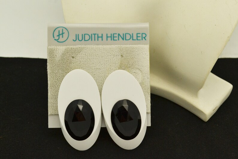 May include: A pair of white oval-shaped earrings with black faceted stones in the center. The earrings are on a white card with the text "Judith Hendler" in teal.