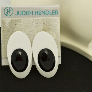 May include: A pair of white oval-shaped earrings with black faceted stones in the center. The earrings are on a white card with the text "Judith Hendler" in teal.