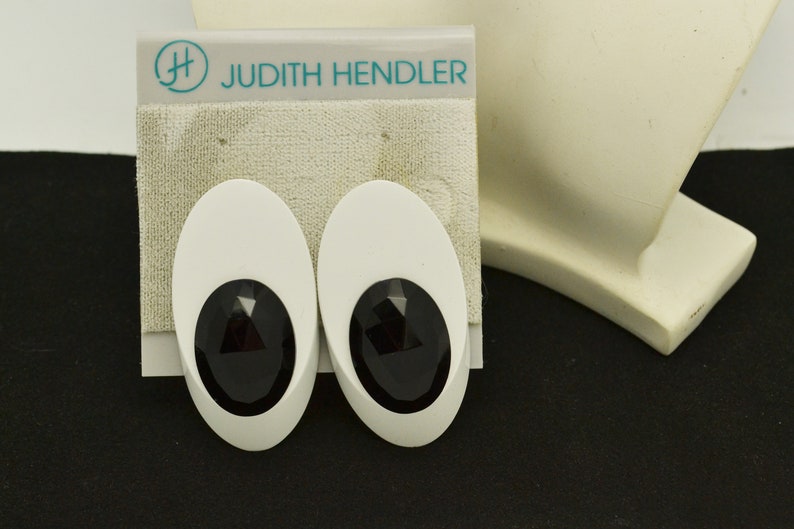 May include: A pair of white oval-shaped earrings with black faceted stones in the center. The earrings are on a white card with the text "Judith Hendler" in teal.