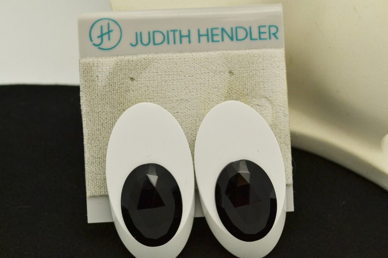May include: A pair of white oval-shaped earrings with black faceted stones. The earrings are on a white card with the text "Judith Hendler" and a logo.