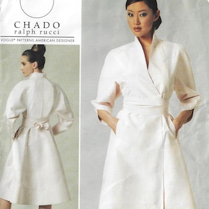 May include: A sewing pattern envelope for a Chado Ralph Rucci Vogue Patterns American Designer coat. The white coat features a wrap-style front, a high collar, three-quarter length sleeves, a waist tie with a bow at the back, and side pockets.
