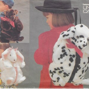 Może przedstawiać: Trzy pluszowe plecaki dla dzieci w kształcie zwierząt. Plecak w kształcie dalmatyńczyka, plecak w kształcie brązowego niedźwiedzia i plecak w kształcie białego królika. Zaprojektowane przez Lindę Carr.