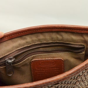 Puede incluir: Vista interior de un bolso tejido marr&oacute;n. El bolso tiene un bolsillo con cremallera, una etiqueta de cuero con texto y ribetes de cuero marr&oacute;n. El interior est&aacute; forrado con una tela beige. El exterior tejido del bolso es visible en la parte inferior.