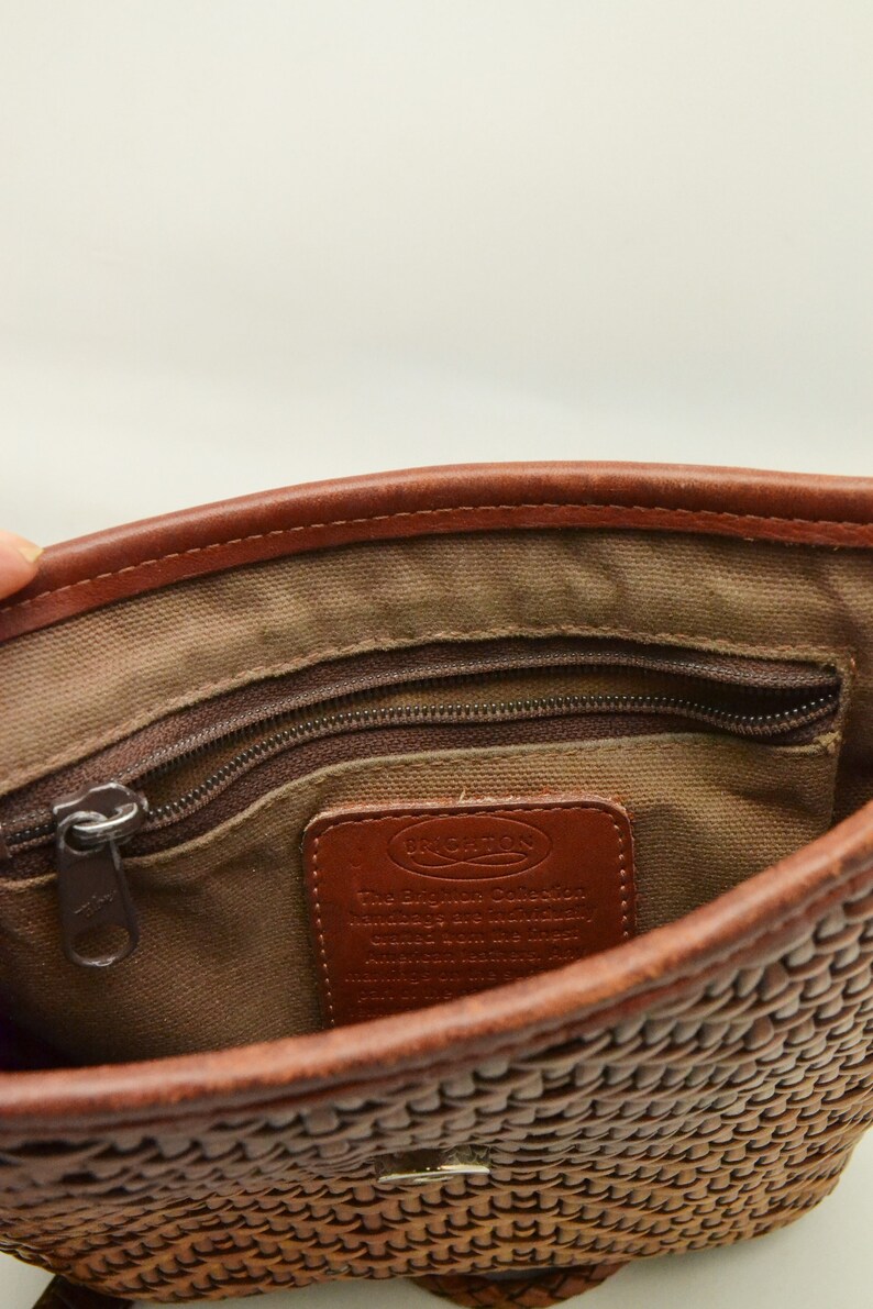 Puede incluir: Vista interior de un bolso tejido marr&oacute;n. El bolso tiene un bolsillo con cremallera, una etiqueta de cuero con texto y ribetes de cuero marr&oacute;n. El interior est&aacute; forrado con una tela beige. El exterior tejido del bolso es visible en la parte inferior.