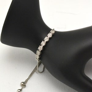 Op de afbeelding: Een zilveren armband met een rij ronde, heldere stenen. De armband heeft een ketting met twee kleine zilveren balletjes aan het einde.