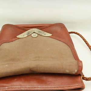 Puede incluir: Mochila de cuero marr&oacute;n con cierre de solapa y correa trenzada. La bolsa presenta un interior de lona beige en contraste y un cierre met&aacute;lico decorativo. El cuero tiene un tono c&aacute;lido y rico, sugiriendo un estilo vintage.