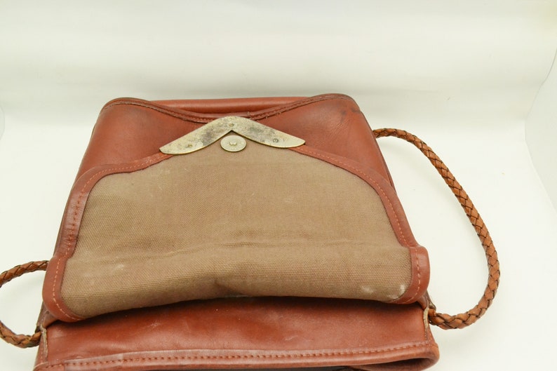 Puede incluir: Mochila de cuero marr&oacute;n con cierre de solapa y correa trenzada. La bolsa presenta un interior de lona beige en contraste y un cierre met&aacute;lico decorativo. El cuero tiene un tono c&aacute;lido y rico, sugiriendo un estilo vintage.