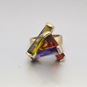 May include: A gold ring with three rectangular gemstones. The gemstones are green, purple, and red.