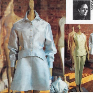 May include: A pale blue women's suit, comprising a collared shirt and a skirt, presented on a mannequin. The suit has a slight shimmer and is fastened with gold-toned buttons. The image also features a portrait of Isabel Toledo.