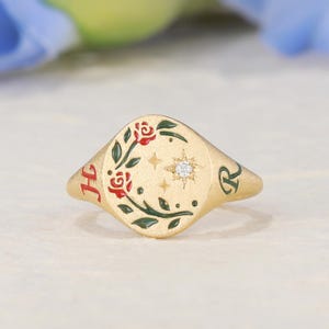 May include: A gold signet ring featuring a matte finish, engraved with the letters H and R on the sides. The face displays a floral motif with red roses, green leaves, and a central starburst design containing a small, clear gemstone.