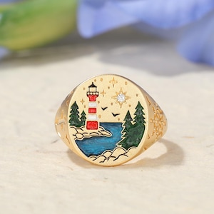 May include: Gold-toned ring with a detailed lighthouse scene. The ring features a red and white lighthouse, green trees, blue water, and a starry sky. The ring has a rounded, oval shape with decorative edges.