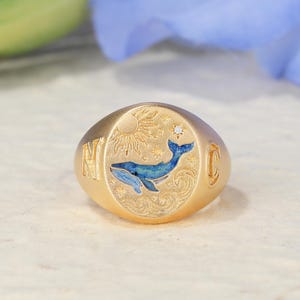 May include: A gold signet ring featuring an engraved sun, stars, and a blue enamel whale swimming above waves. The letter N is engraved on the left side of the band, and the letter C is engraved on the right side. A small clear stone is set near the stars.