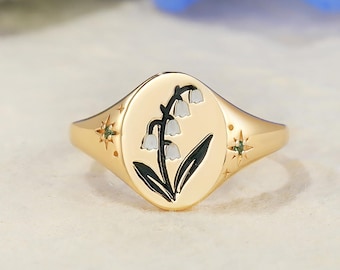 Puede incluir: Un anillo de sello dorado con una cara ovalada que presenta un diseño grabado de lirio de los valles en blanco y negro. La banda del anillo está decorada a ambos lados de la cara ovalada con un pequeño motivo de estrella de cuatro puntas.