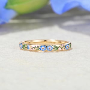 May include: A gold band ring featuring a continuous enamel pattern of blue forget-me-not flowers with green leaves and small pink buds. The metal surface is polished and reflective, showcasing a delicate floral design wrapped around the entire circumference.