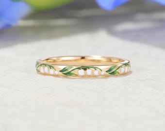 Puede incluir: Un anillo de banda dorada que presenta un patrón de esmalte continuo de flores blancas de lirio de los valles con hojas verdes que envuelven toda la circunferencia de la superficie metálica, mostrando un diseño botánico detallado y pulido.
