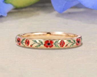 Puede incluir: Un anillo de banda fina de oro con un patrón de esmalte continuo de flores de amapola rojas con tallos y hojas verdes. El metal tiene un acabado pulido y el diseño floral envuelve toda la circunferencia de la pieza de joyería circular.