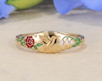 May include: A gold-toned ring featuring an engraved hummingbird at the center, flanked by a red enamel rose with green leaves on one side and white lily of the valley flowers with green leaves on the other, accented by small star engravings.