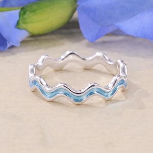 May include: A silver ring featuring a continuous wavy, scalloped design. The band has a polished metallic finish with a thin, light blue enamel inlay running along the centre of the wave pattern, creating a smooth and consistent ripple effect.