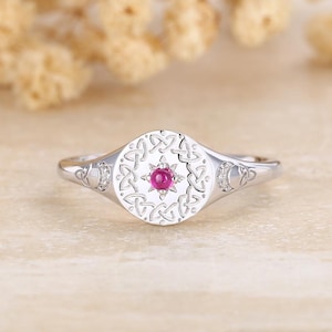 May include: A silver ring with a circular face featuring a central red gemstone surrounded by a Celtic knot design. The ring has a polished finish and small accent stones on each side of the face.