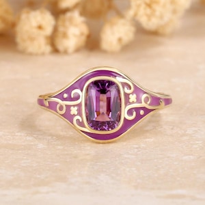 May include: A gold ring with a rectangular, deep purple gemstone set in a light purple enamel setting. The enamel features decorative swirls and accents. The ring is set on a neutral surface.