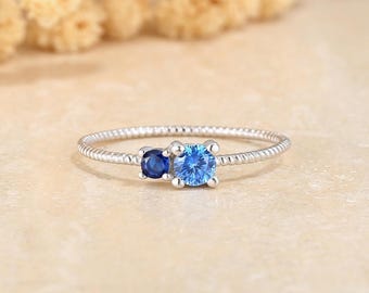 May include: A silver ring with a textured band, featuring two blue gemstones. One gemstone is a smaller, dark blue, round stone, and the other is a larger, light blue, round stone. The ring is set against a soft, neutral background.