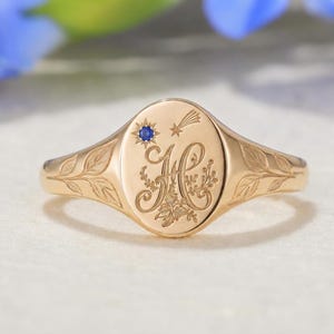 May include: A gold signet ring featuring an oval face with an engraved letter H, a small blue gemstone set in a starburst, and a shooting star motif. The band is decorated with engraved leaf patterns extending from the central oval face.