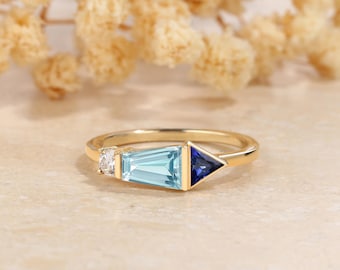 May include: A gold ring featuring a rectangular light blue gemstone, a small white gemstone, and a triangular dark blue gemstone. The ring is set against a neutral background with blurred floral accents.