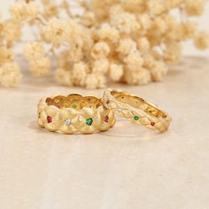 May include: Two gold rings with textured surfaces. One ring is wider, the other is thinner. Both are adorned with alternating red, green, and clear gemstones. The rings are set against a soft, neutral background with dried flowers.